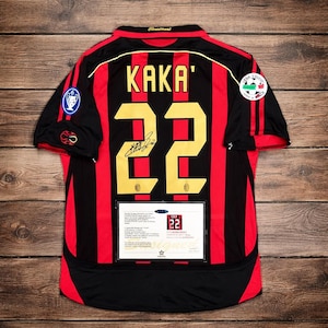 May include: Black and red football shirt with the name "KAKA'" and the number "22" in gold. The shirt features a signature and patches. A framed certificate is displayed below the number.