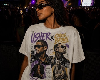 Puede incluir: Una camiseta blanca de manga corta con un estampado gráfico de Usher y Chris Brown. El diseño incluye texto en morado y dorado, retratos de ambos artistas con gafas de sol e imágenes más pequeñas de sus actuaciones y canciones.