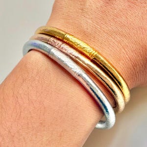 Pode incluir: Três pulseiras metálicas em ouro, ouro rosa e prata são empilhadas em um pulso. A pulseira dourada tem um design texturizado. As pulseiras são lisas e brilhantes, refletindo a luz. A joia é simples e elegante.