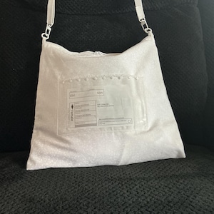 May include: A white crossbody bag with a subtle tonal pattern and a clear plastic pocket on the front. The pocket contains a printed document with the text Elin, son, and Female, along with various dates and administrative information.