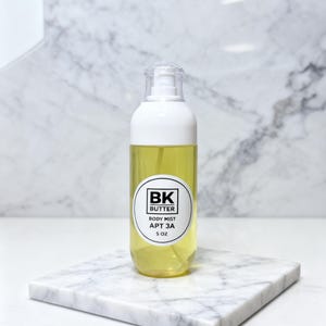 May include: A clear body mist bottle with a white top and a yellow liquid inside. The bottle has a label that reads "BK BUTTER BODY MIST APT 3A 5 oz." The bottle is on a white marble square.