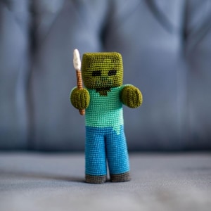 Puede incluir: Una figura de zombi de Minecraft hecha a mano con ganchillo. La figura tiene una cabeza verde, una camisa turquesa y pantalones azules. Sostiene una pequeña lanza con punta blanca. La figura está sobre una superficie gris.