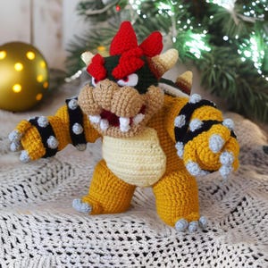 Könnte beinhalten: Gehäkelte Bowser-Figur mit gelbem Körper, schwarzen Armbändern mit Stacheln und einer roten, stacheligen Krone. Die Figur hat eine beige Schnauze, weiße Zähne und rote Augen. Sie steht auf einer weißen, strukturierten Oberfläche, mit verschwommenem Hintergrund eines Weihnachtsbaums und einer goldenen Kugel.