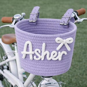以下が含まれることがあります： 白い文字で「Asher」と書かれた名前と白いリボンが付いた、ラベンダー色の自転車用バスケット。バスケットは、ラベンダー色のストラップとシルバーのバックルでハンドルバーに取り付けられています。自転車には茶色のハンドルバーと茶色のシートが付いています。