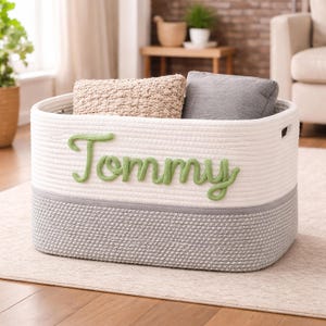 Op de afbeelding: Een rechthoekige opbergmand van gewikkeld wit en grijs touw met geïntegreerde handgrepen aan de zijkanten. Op de voorzijde staat de naam Tommy geborduurd in een lichtgroen cursief lettertype. In de mand liggen een beige en een grijs kussen.