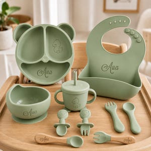 Puede incluir: Un juego de alimentación de silicona en color verde salvia que incluye un plato dividido con forma de oso, un babero con cuatro estrellas recortadas, un cuenco, un vaso con tapa y pajita, y varios cubiertos. Cada pieza lleva el nombre Aua y un dibujo.