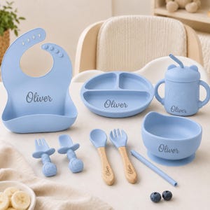 May include: A light blue silicone baby feeding set featuring the name Oliver printed on each piece. The set includes a bib with a crumb catcher, a three-section divided plate, a lidded cup with two handles and a straw, a bowl, and four utensils.