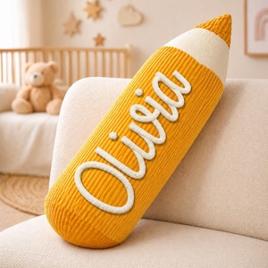 May include: A yellow pencil-shaped pillow with the name "Olivia" in white script. The pillow has a corduroy texture and a white tip. It is resting on a cream-colored sofa.