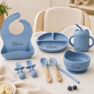 May include: A blue silicone feeding set featuring a bib, a three-section plate, a lidded cup with a straw, a bowl, and two sets of utensils. Each item is personalized with the name Oliver. The utensils include silicone and wooden handles.