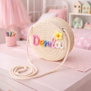 May include: A circular straw handbag with a long, braided strap. The bag is decorated with the name "Denla" in colourful, embroidered letters and a yellow flower with an orange centre. It rests on a pink surface.