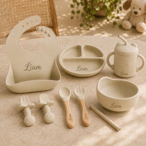 May include: A beige silicone baby feeding set featuring a bib, a three-section plate, a lidded cup with handles and a straw, a bowl, and two sets of utensils. Each item is personalized with the name Liam in a cursive script font.