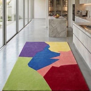 Op de afbeelding: Een rechthoekig vloerkleed met een modern geometrisch ontwerp. Het kleed heeft grote, onregelmatige vormen in levendige kleuren: groen, paars, blauw, geel, roze en rood. Het kleed ligt op een grijze betonnen vloer in een moderne binnenruimte.
