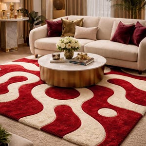 Puede incluir: Salón con sofá crema y cojines burdeos y dorados. Una mesa de centro redonda con tapa de mármol y base dorada se encuentra sobre una alfombra estampada roja y crema. Una lámpara de araña cuelga encima.