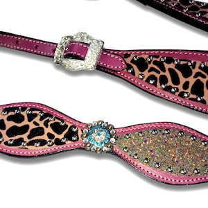 May include: Close-up of decorative horse tack. The tack features a pink leather base with a leopard print design and silver-colored studs. One piece has a silver buckle, and another has a blue and clear jewel-encrusted centerpiece.
