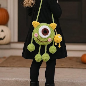May include: A round, green, crocheted monster bag featuring a single large central eye with a dark brown pupil, two small yellow ears, pink cheeks, and a wide brown smile. The bag has four dangling green pom-pom limbs and a small yellow pom-pom charm.