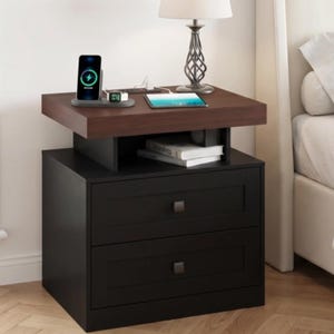 May include: A two-drawer nightstand with a dark brown top surface and a black base. The top features a wireless charging station with a smartphone and smartwatch, a tablet, and a lamp. An open shelf below the top holds two books.