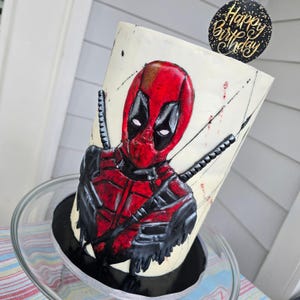 May include: A cylindrical birthday cake featuring a detailed depiction of Deadpool in red and black. The cake has a white base with black accents and two katana swords. A black and gold "Happy Birthday" topper is placed on top.