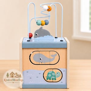 May include: A wooden activity cube featuring a wire bead maze on top with colorful wooden beads and a dolphin cutout. The front panel displays sliding puzzle blocks depicting a whale and a sea turtle on a light blue and natural wood background.