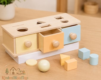 May include: A wooden shape sorting box with three drawers in pastel yellow, orange, and blue. The top panel features cutouts for a circle, triangle, rectangle, square, and line. Includes matching wooden shapes: a sphere, cylinder, cube, and prism.