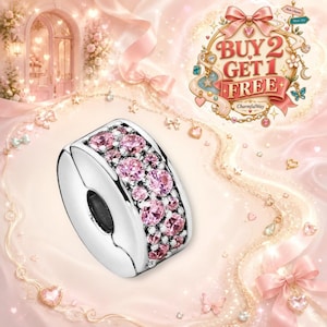 May include: A silver charm bead featuring a central band encrusted with multiple round, faceted pink stones. The bead has a smooth, polished metallic finish on the sides and a circular opening in the center for threading onto a bracelet chain.