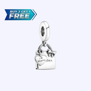 Può includere: Un charm d'argento con un design a forma di borsa della spesa. Il charm presenta un'etichetta a forma di cuore con le parole "I love shopping" e un fiocco. Sulla borsa della spesa è stampata la parola "ORA".