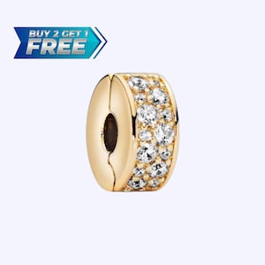 Pode incluir: Berloque dourado com uma fileira de pedras claras e brilhantes. O berloque tem um design de fecho e é de formato oval. A imagem inclui o texto "BUY 2 GET 1 FREE" em uma faixa azul e branca.