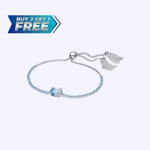 Puede incluir: Una pulsera plateada con una fila de pequeñas piedras azul claro y una piedra azul ovalada más grande en el centro. La pulsera tiene un cierre deslizante y dos borlas decorativas. La imagen también incluye un gráfico promocional azul y blanco que dice "BUY 2 GET 1 FREE".