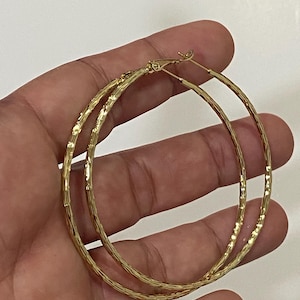 May include: A pair of gold-colored hoop earrings. The earrings have a textured surface and a classic circular design. The hoops are held in a hand against a white background. The earrings are a simple yet elegant accessory.