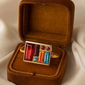 May include: A gold-toned ring with a rectangular face, set with colorful rectangular and round gemstones in red, brown, orange, blue, and turquoise. The ring is displayed in an open, brown velvet jewelry box.