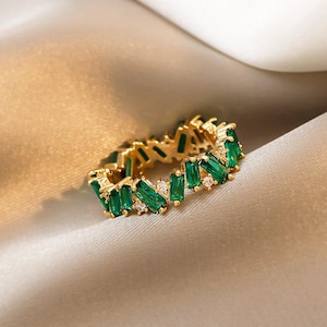 May include: A gold ring set with emerald-green baguette-cut gemstones and small, clear accent stones. The ring's design is a continuous band of alternating green and clear stones, creating a sparkling, elegant look. The ring is set against a soft, neutral background.