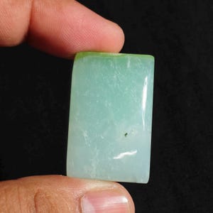 May include: A rectangular, polished gemstone cabochon with a smooth, flat surface. The stone is a pale, translucent mint green colour with subtle white cloudy inclusions and a single tiny dark speck visible near the centre of the piece.