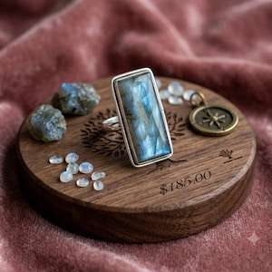 May include: A rectangular labradorite ring with a silver setting, displayed on a circular wooden base alongside two raw labradorite stones, several small moonstone cabochons, and a small bronze-toned compass charm with the text $185.00.