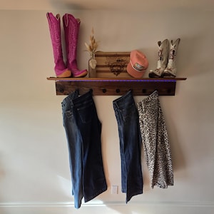 May include: A wooden shelf with a row of hooks, displaying pink cowboy boots, a wooden cutting board, a pink cap, and white cowboy boots. Below, two pairs of jeans and leopard print trousers hang from the hooks. The shelf has a decorative light.