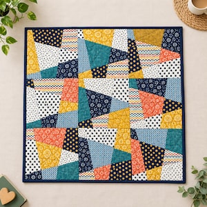 May include: A square fabric quilt featuring a patchwork design of irregular geometric shapes. The pattern includes various prints in navy blue, mustard yellow, teal, white, and coral, with polka dots, floral motifs, and chevron stripes.