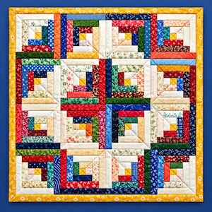 Op de afbeelding: Een vierkante quilt met een log cabin-patroon bestaande uit zestien blokken. Elk blok toont een verscheidenheid aan kleurrijke bloemrijke en geometrische stoffen stroken in rood, blauw, groen en geel, omlijst door een gele rand met een wit madeliefjespatroon.