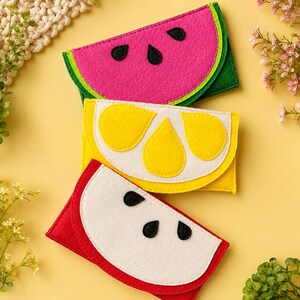 Op de afbeelding: Drie vilten etuis in de vorm van fruitpartjes. Het bovenste etui is een roze watermeloenpartje met zwarte pitjes en een groene schil. Het middelste etui is een geel citroenpartje met witte segmenten. Het onderste etui is een rood appelpartje met wit vruchtvlees en zwarte pitjes.