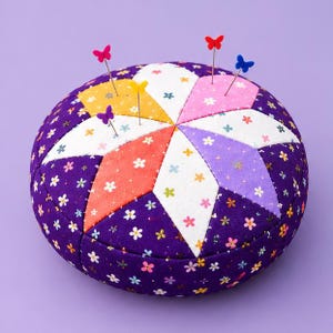 May include: A round fabric pincushion featuring a purple base with a multi-colored star-shaped patchwork pattern of white, yellow, orange, pink, and lavender floral fabric. Four pins with colorful butterfly-shaped heads are inserted.