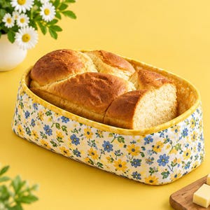 May include: An oval fabric bread warmer basket featuring a white exterior with a pattern of small yellow and blue flowers and green leaves, lined with a yellow and white floral fabric, containing a loaf of golden brown baked bread.