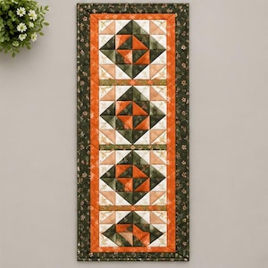May include: A rectangular quilted table runner featuring four repeating square blocks in a geometric pattern. The design uses shades of dark green, burnt orange, and cream, with a dark green floral border and a solid orange inner frame.