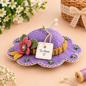 May include: A purple felt pincushion shaped like a flower with a scalloped edge, decorated with a pink fabric flower, green leaves, and small green beads. A white square tag attached with a string reads To: Mom with a heart symbol.
