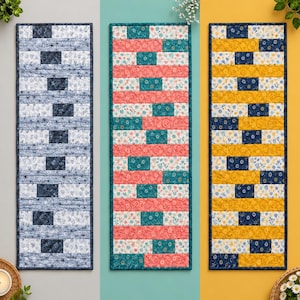 May include: Three rectangular quilted table runners featuring a brick-like pattern. The left runner is blue and white, the centre runner is coral and teal with floral motifs, and the right runner is yellow and navy blue with small floral patterns.