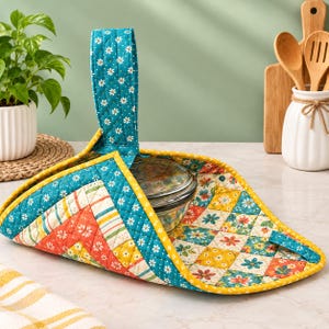 May include: A quilted fabric casserole carrier featuring a teal background with a white daisy pattern, accented by orange and yellow geometric floral patches. The carrier includes a single upright handle and a yellow fabric trim border.