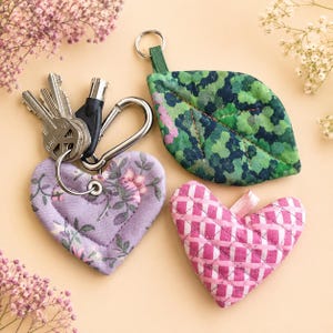 May include: Three quilted fabric keychains: one heart-shaped with a lavender floral print attached to a metal keyring with keys, one leaf-shaped with a green mottled pattern, and one heart-shaped with a pink and white checkered pattern.