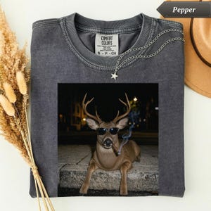 May include: A charcoal gray crew neck t-shirt featuring a graphic print of a deer wearing black sunglasses and holding a lit cigar in its mouth. The shirt has a Comfort Colors label at the inner neckline and a silver star pendant necklace.