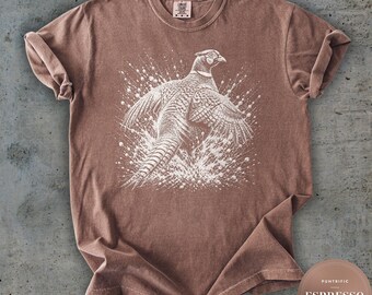 May include: A brown short-sleeve t-shirt featuring a white graphic illustration of a pheasant in flight surrounded by a splatter effect. The shirt has a crew neckline and a label at the inner collar. A circular badge in the corner reads PUNTRIFIC ESPRESSO UNISEX.