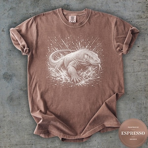 Può includere: Una t-shirt marrone con un'illustrazione grafica bianca di un drago di Komodo circondato da un effetto a spruzzo. La maglietta ha un girocollo e maniche corte. L'etichetta interna sul collo riporta il testo Comfort Colors e la taglia M.