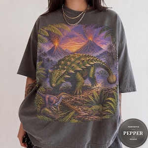 May include: A charcoal gray short-sleeve t-shirt featuring a large graphic print of a prehistoric landscape with an ankylosaurus, a smaller dinosaur, lush green ferns, and two erupting volcanoes under a vibrant orange and purple sunset sky.