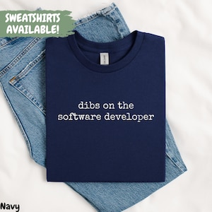May include: A navy blue t-shirt with the white text "dibs on the software developer". The t-shirt is laid on blue denim jeans. A green banner in the top left corner reads "SWEATSHIRTS AVAILABLE!"