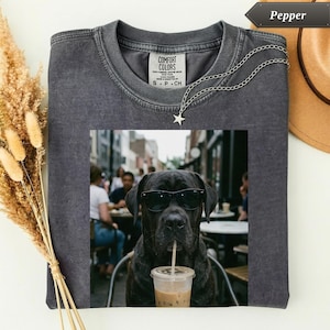 May include: A charcoal grey crew neck t-shirt featuring a graphic print of a black dog wearing black sunglasses and drinking from a clear plastic cup with a straw. The shirt has a Comfort Colors label at the neck and a silver star pendant necklace.