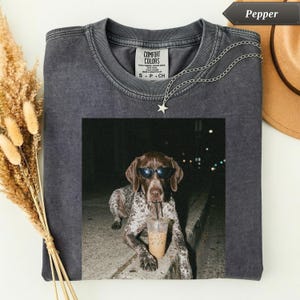 May include: A dark gray Comfort Colors t-shirt with a photo of a dog wearing sunglasses and drinking from a cup. A silver chain with a star pendant is draped on the shirt. The word "Pepper" is in a banner at the top.
